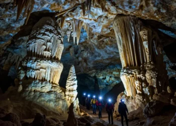 A dramatic, photorealistic image of an underground cave in North America, massive stalactites and stalagmites, crystal formations glowing with soft blue and amber light, a small group of explorers with headlamps walking on a narrow path, deep darkness surrounding, high detail, cinematic lighting, 8K resolution.